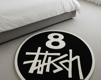 May include: A round black and white rug with the number 8 and a stylized logo. The rug has a white border and is placed on a gray carpeted floor. The rug is in a bedroom setting.