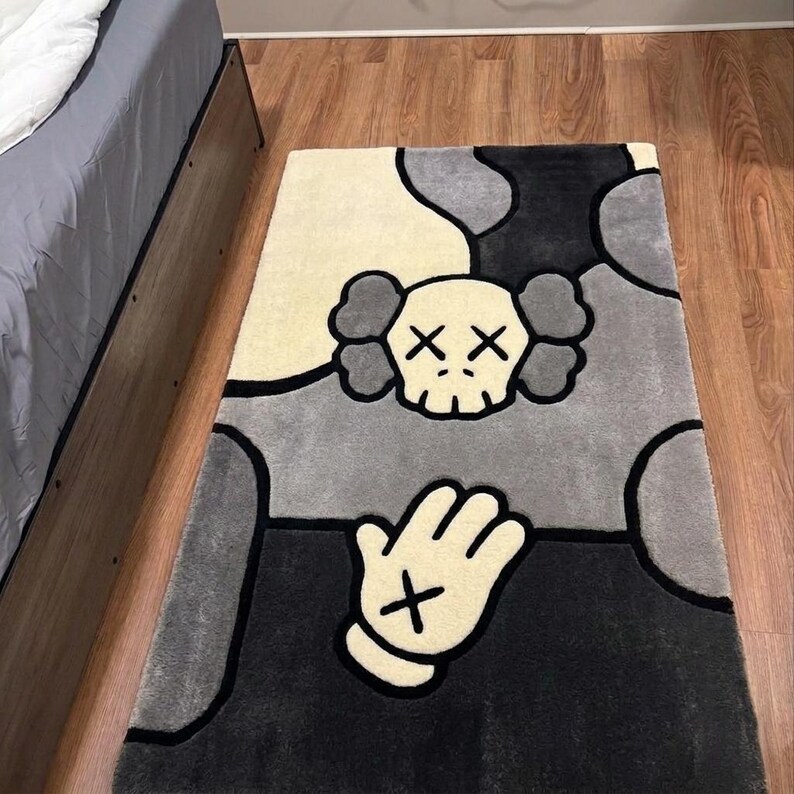 K&ouml;nnte beinhalten: Ein rechteckiger Teppich mit einem cremefarbenen Totenkopf mit X-f&ouml;rmigen Augen und einer passenden cremefarbenen Hand mit einem X auf der Handfl&auml;che. Der Teppich hat ein segmentiertes Hintergrundmuster in Schwarz, Dunkelgrau und Hellgrau mit dicken schwarzen Umrandungen.