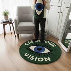 May include: A green round rug with a large eye design and the words "CATCH OUR VISION" in white. A rolled-up matching rug is held by a person. The rug has a blue iris and white eye. The floor is wood.