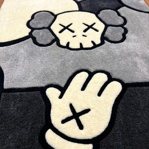 K&ouml;nnte beinhalten: Ein rechteckiger getufteter Teppich mit einem stilisierten Totenkopf-Charakter mit kreuzf&ouml;rmigen Augen und wolkenartigen Ohren in Creme- und Graut&ouml;nen. Unter dem Totenkopf ist eine gro&szlig;e cremefarbene Cartoon-Hand mit einem schwarzen Kreuz abgebildet.