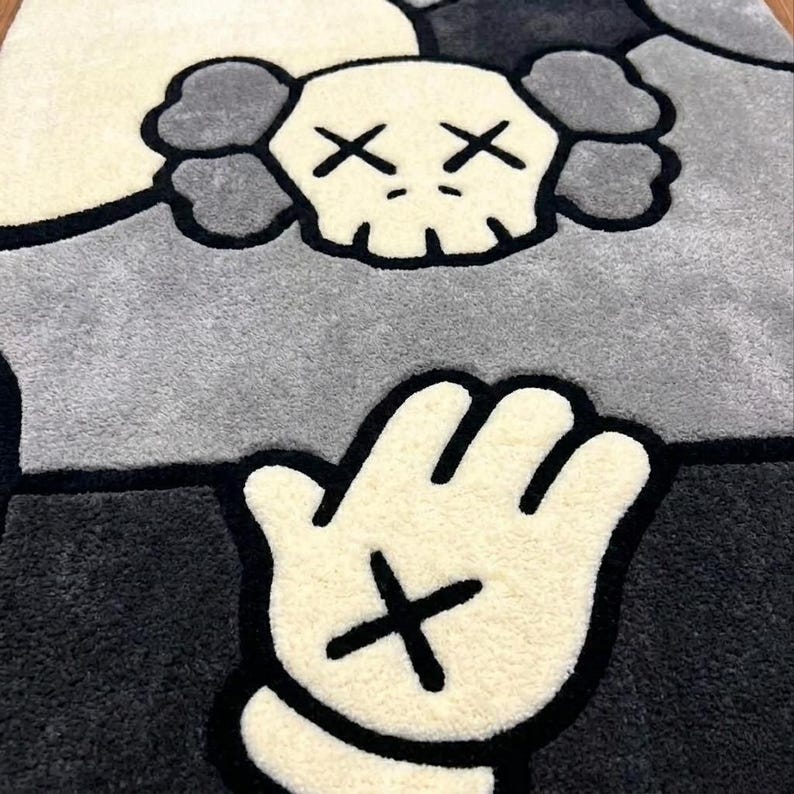 K&ouml;nnte beinhalten: Ein rechteckiger getufteter Teppich mit einem stilisierten Totenkopf-Charakter mit kreuzf&ouml;rmigen Augen und wolkenartigen Ohren in Creme- und Graut&ouml;nen. Unter dem Totenkopf ist eine gro&szlig;e cremefarbene Cartoon-Hand mit einem schwarzen Kreuz abgebildet.