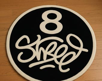 May include: A round black rug with a cream-colored border. The rug features the number "8" and the word "Shred" in a cream-colored, stylized font. The rug is placed on a wooden floor.