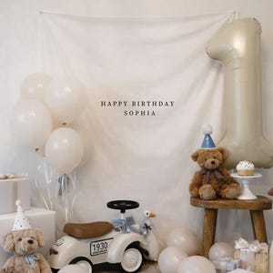 Può includere: Una scena di compleanno con palloncini bianchi, regali e un grande palloncino con il numero uno. Sono presenti un orsacchiotto e un'auto giocattolo. Il testo "HAPPY BIRTHDAY SOPHIA" è visualizzato sullo sfondo. Sono presenti anche un cupcake e un'oca.