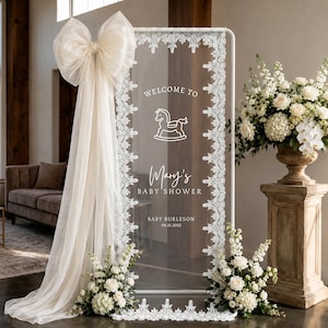 Puede incluir: Un letrero rectangular de acrílico transparente en un marco blanco, decorado con encaje blanco y un gran lazo de tela color crema. El texto blanco dice Welcome to Mary's Baby Shower, Baby Burleson, 09.21.2025, e incluye un icono de caballo balancín.
