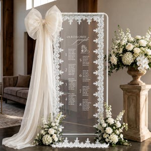 May include: A tall, rectangular clear acrylic seating chart sign framed in white with intricate white lace trim. The sign features the text "OUR FAVORITE people" and lists names under eight numbered tables. A large, sheer cream-colored fabric bow is attached to the top left corner.