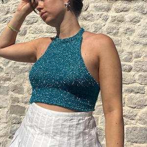 Pode incluir: Top halter turquesa com um aspeto brilhante e texturizado. É combinado com uma saia plissada branca. O design do top apresenta um decote alto e um comprimento curto, adequado para climas quentes ou para sobreposição.