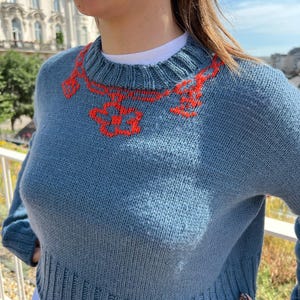 Puede incluir: Un suéter de punto azul de manga larga con cuello redondo y dobladillo acanalados. La parte superior del pecho está decorada con un patrón bordado de cadena de eslabones naranja que incluye un dije en forma de flor en el centro.