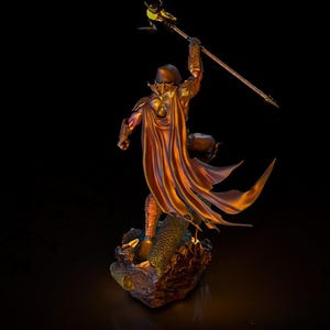 Puede incluir: Estatua detallada de una figura de guerrero en una pose din&aacute;mica, sosteniendo una lanza en alto. La figura est&aacute; vestida con una armadura ornamentada y una capa fluida, con una combinaci&oacute;n de colores bronce, oro y p&uacute;rpura. La estatua descansa sobre una base texturizada.