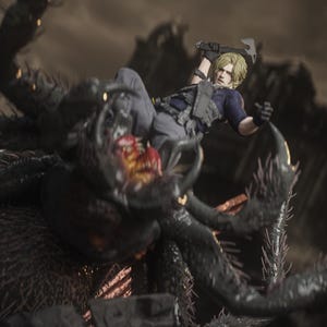 May include: A detailed action figure of a character with blonde hair, wearing a blue shirt and tactical vest, wielding a hatchet. The figure is posed mid-action, battling a large, monstrous spider with a menacing open mouth. The scene is set against a blurred, dark background.