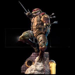 以下が含まれることがあります： ミュータント・タートルズのラファエルの詳細な像。フィギュアは緑色で赤いマスクをしており、茶色の鎧を着てサイを持っています。矢が背中に埋め込まれています。像はレンガと石の台座に立っています。