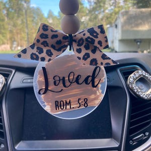May include: A car air freshener with a clear, round pendant featuring the word "Loved" and the text "ROM. 5:8" in black script. It has a brown and black leopard print bow and wooden beads.
