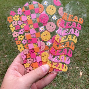 May include: A hand holding a collection of clear acrylic bookmarks. The bookmarks feature colorful designs including flowers, checkerboard patterns, smiley faces, and the word "READ" in a retro font. Each bookmark has a hole at the top.