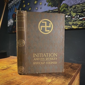 May include: A vintage book titled "Initiation and Its Results" by Rudolf Steiner. The cover is brown with gold lettering and a gold swastika symbol. The book is standing on a wooden surface.