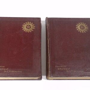May include: Two antique books with burgundy covers. Each book features a gold sunburst emblem and the text "Editoriship of Dr. L.W. deLaurence". The books are presented against a white backdrop.