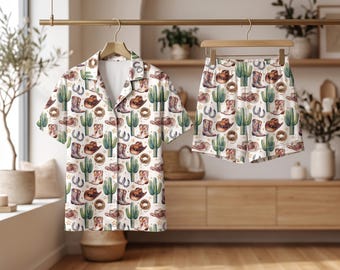 May include: A two-piece pajama set consisting of a short-sleeved button-down shirt and matching shorts. The fabric is white and features a repeating pattern of brown cowboy hats, brown cowboy boots, green cacti, and brown coiled ropes.