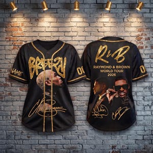 May include: A black baseball jersey with gold trim and glittery gold text reading BREEZY on the front. The back features the text R&B RAYMOND & BROWN WORLD TOUR 2026, images of two men, and gold signatures. The jersey has short sleeves and buttons.
