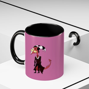 May include: A ceramic mug with a black handle and interior, featuring a cartoon character on a pink background. The character is red with yellow eyes, black and white horns, and a black suit. The mug is on a white surface.