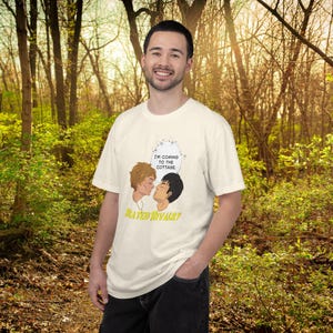 May include: Cream-colored t-shirt featuring a cartoon illustration of two men kissing, with the text "I'M COMING TO THE COTTAGE" in a speech bubble. Below the illustration, the text "FATED RIVALRY" is displayed in yellow.