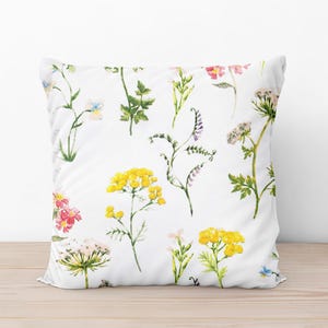Op de afbeelding: Wit vierkant kussen met een aquarel bloemenpatroon. Het ontwerp toont verschillende wilde bloemen in geel, roze, paars en groen. Het kussen ligt op een licht houten oppervlak.