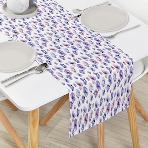 Pode incluir: Um caminho de mesa branco com um padrão repetido de formas florais roxas e azuis. O caminho de mesa está sobre uma mesa branca com pratos, talheres e uma xícara pequena.