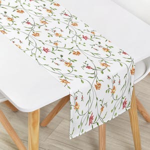 Könnte beinhalten: Weißer Tischläufer mit floralem Muster. Der Läufer zeigt ein sich wiederholendes Muster aus zarten Ranken, die mit gelben, orangefarbenen und roten Blüten und grünen Blättern verziert sind. Er liegt auf einem weißen Tisch und hebt das florale Muster hervor.