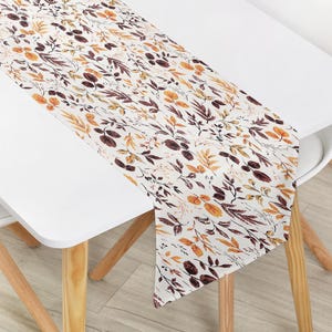 May include: A rectangular table runner featuring a white background with a repeating pattern of autumn-themed leaves in shades of orange, yellow, and dark brown. The botanical print includes various leaf shapes and stems arranged across the fabric.