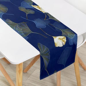 Pode incluir: Um caminho de mesa retangular com um fundo azul-marinho profundo decorado com um padrão repetitivo de folhas de ginkgo biloba. As folhas são apresentadas em tons de dourado, branco e azul-claro com detalhes de linhas finas e delicadas.