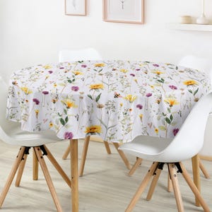 Peut inclure: Une nappe ronde à fond blanc ornée d'un motif botanique composé de tournesols jaunes, de fleurs sauvages violettes, de feuillages verts et de petites abeilles, recouvrant une table ronde en bois entourée de quatre chaises blanches.