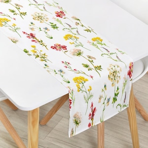 Peut inclure: Chemin de table blanc avec un motif floral de fleurs sauvages jaunes, rouges et violettes. Le chemin de table est drapé sur une table blanche, mettant en valeur le motif botanique coloré. La table a des pieds en bois clair.