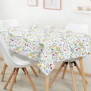 Könnte beinhalten: Ein rechteckiges weißes Tischtuch mit einem farbenfrohen Blumenmuster, darunter rote, gelbe und blaue Blumen, bedeckt einen Tisch. Das Tischtuch liegt auf einem Tisch mit weißen Stühlen und Holzbeinen. Das Thema ist Frühling oder Sommer.