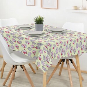 Op de afbeelding: Een rechthoekig tafelkleed met een lichtgele achtergrond en een herhalend patroon van cupcakes in roze, grijze en witte tinten. Het tafelkleed ligt over een tafel met vier witte stoelen en een kleine plant in een pot.