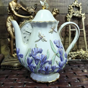 Könnte beinhalten: Eine weiße Keramik-Teekanne mit Deckel, verziert mit einem Schmetterling und Libellen. Die Teekanne ist mit lila Irisblüten und goldenen Verzierungen versehen. Sie hat einen gebogenen Griff und einen Ausguss.