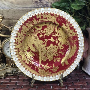 May include: A round ceramic plate featuring a deep red background with intricate gold illustrations of two large birds, floral patterns, and a butterfly. The rim is decorated with a white and gold lattice border with small floral accents.