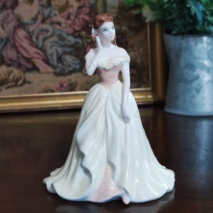 May include: A porcelain figurine of a woman in a long white gown with a pink bodice and accents. The woman has auburn hair and is posed with her hand near her face. The figurine is a decorative item, likely for display.