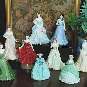 May include: A collection of porcelain figurines depicting women in elegant gowns. The figurines are in various colors, including green, red, blue, and white. The dresses feature intricate details, and the figurines are posed in graceful stances.