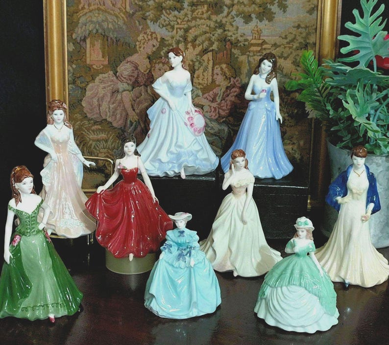 May include: A collection of porcelain figurines depicting women in elegant gowns. The figurines are in various colors, including green, red, blue, and white. The dresses feature intricate details, and the figurines are posed in graceful stances.