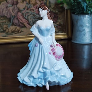 May include: A porcelain figurine of a woman in a light blue dress, holding a pink-trimmed hat. The dress has ruffled layers and pink floral accents. The woman has brown hair and is looking to the side. The figurine is on a dark wooden surface.