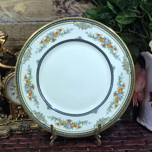 May include: A round ceramic dinner plate with a cream-coloured centre and a wide border featuring a repeating pattern of orange and blue flowers, green foliage, and gold scrollwork accents, finished with a thin gold rim and a dark blue inner band.