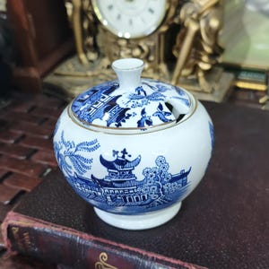Puede incluir: Un azucarero redondo de cerámica blanca y azul con tapa. El azucarero presenta un diseño Willow con ribete dorado. La tapa tiene un pequeño asa y una ranura para una cuchara. El azucarero es redondo y se asienta sobre una base pequeña.