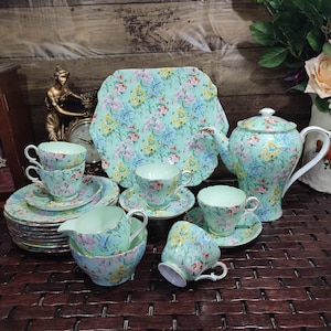 Könnte beinhalten: Ein Vintage-Teeservice mit floralem Muster in Blau, Gelb und Rosa auf mintgrünem Hintergrund. Das Set umfasst eine Teekanne, Tassen, Untertassen, einen Servierteller und einen Sahnekännchen. Die Ränder sind goldfarben.