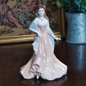 May include: A porcelain figurine of a woman in a long, flowing pink gown with a light blue shawl. The figurine has brown hair and is adorned with a gold necklace and bracelet. The dress features white floral accents.