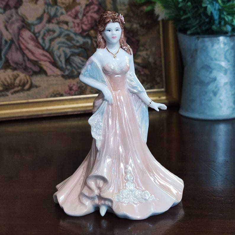 May include: A porcelain figurine of a woman in a long, flowing pink gown with a light blue shawl. The figurine has brown hair and is adorned with a gold necklace and bracelet. The dress features white floral accents.