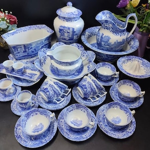 Puede incluir: Un conjunto de vajilla de porcelana azul y blanca, que incluye tazas, platillos, una jarra, un cuenco y fuentes. Las piezas presentan un diseño paisajístico. El conjunto está dispuesto sobre una superficie oscura.