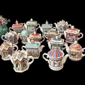 Può includere: Una collezione di quattordici piccole teiere in ceramica, ognuna caratterizzata da illustrazioni colorate uniche di figure e scene storiche, con manici e coperchi rifiniti in oro. Le teiere presentano forme e design differenti.