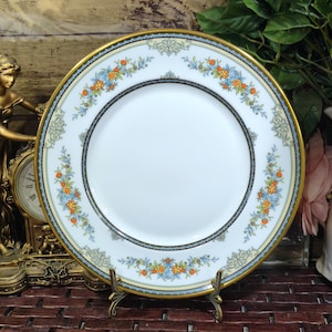 May include: A round white porcelain dinner plate featuring a gold rim and a decorative border pattern of small orange and blue flowers with green foliage. The plate has a central white circular area and is displayed on a small metal stand.