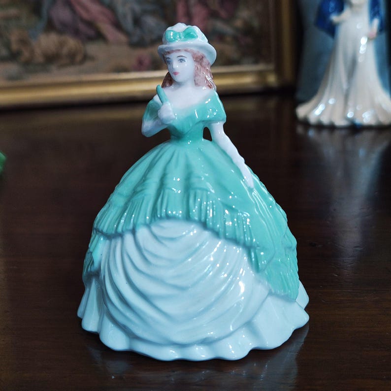 May include: A porcelain figurine of a woman in a teal dress and hat. The figurine has a light blue skirt and a teal bodice with white gloves. The woman is holding a small object in her right hand. The figurine is a decorative item.
