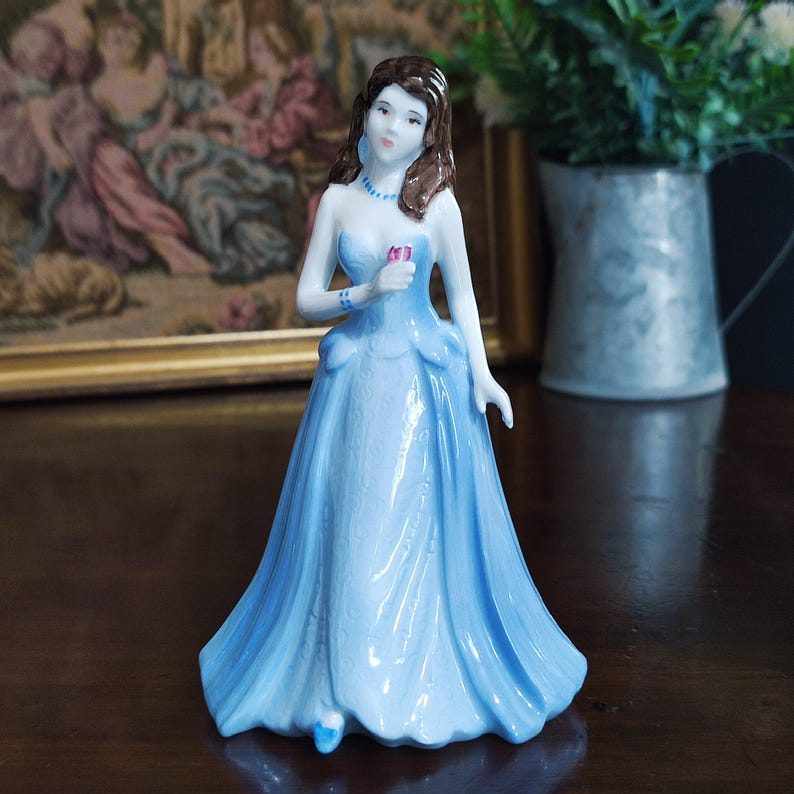 May include: A porcelain figurine of a woman in a light blue strapless gown, holding a pink flower. The figurine has brown hair and is adorned with blue jewelry. The dress features a flowing skirt and detailed bodice. A classic collectible.