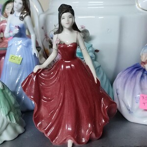 May include: A porcelain figurine of a woman in a flowing red dress. The figurine has dark hair and is adorned with a small pink flower. Other figurines in blue and purple dresses are visible in the background.
