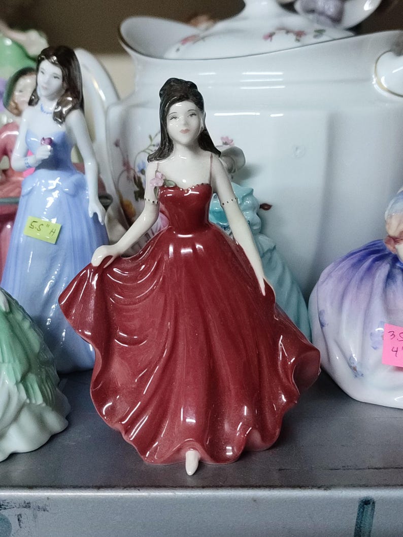 May include: A porcelain figurine of a woman in a flowing red dress. The figurine has dark hair and is adorned with a small pink flower. Other figurines in blue and purple dresses are visible in the background.