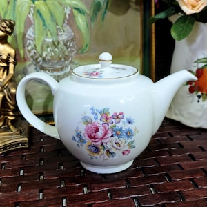 Puede incluir: Tetera de porcelana blanca con un diseño floral de flores rosas, azules y amarillas. La tetera tiene un asa curva, un pico y una tapa con un pomo dorado. Un diseño clásico para servir té.